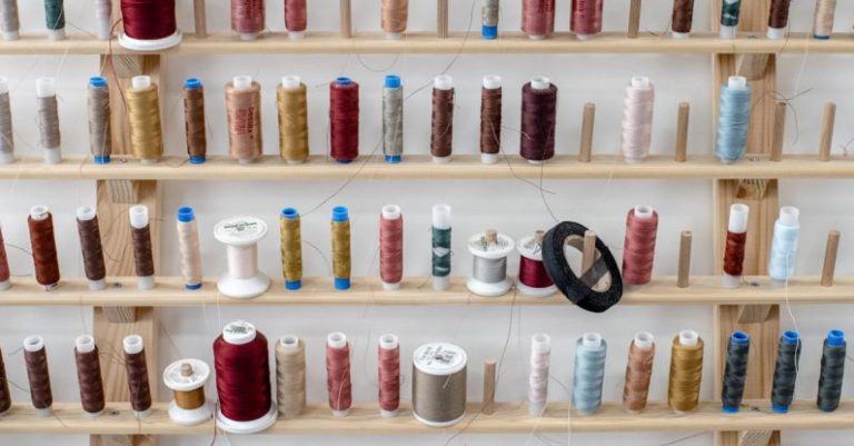 Slip Stitch - Stand with assorted colorful spools of thread for sewing placed near white wall inside dressmaker workshop or tailor atelier for fashion design and apparel creation